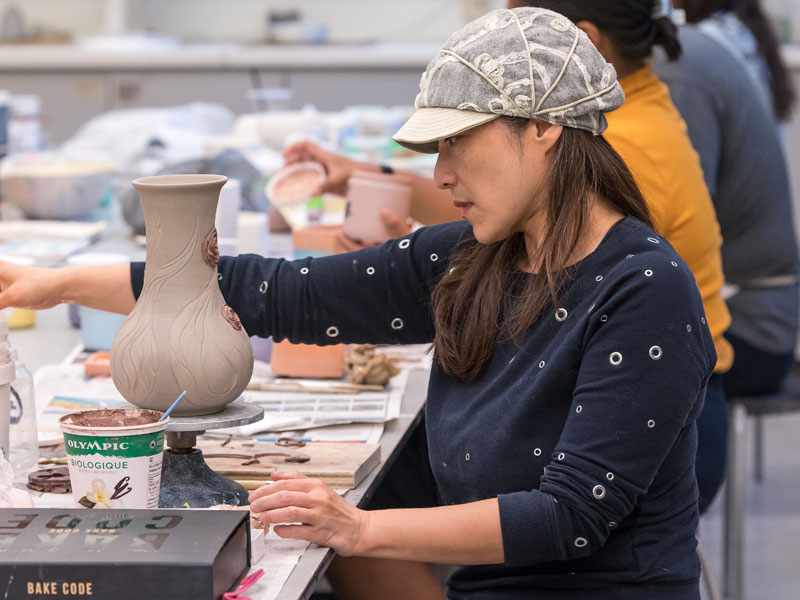 A woman sitting at a table working on a clay vase with other people in the background