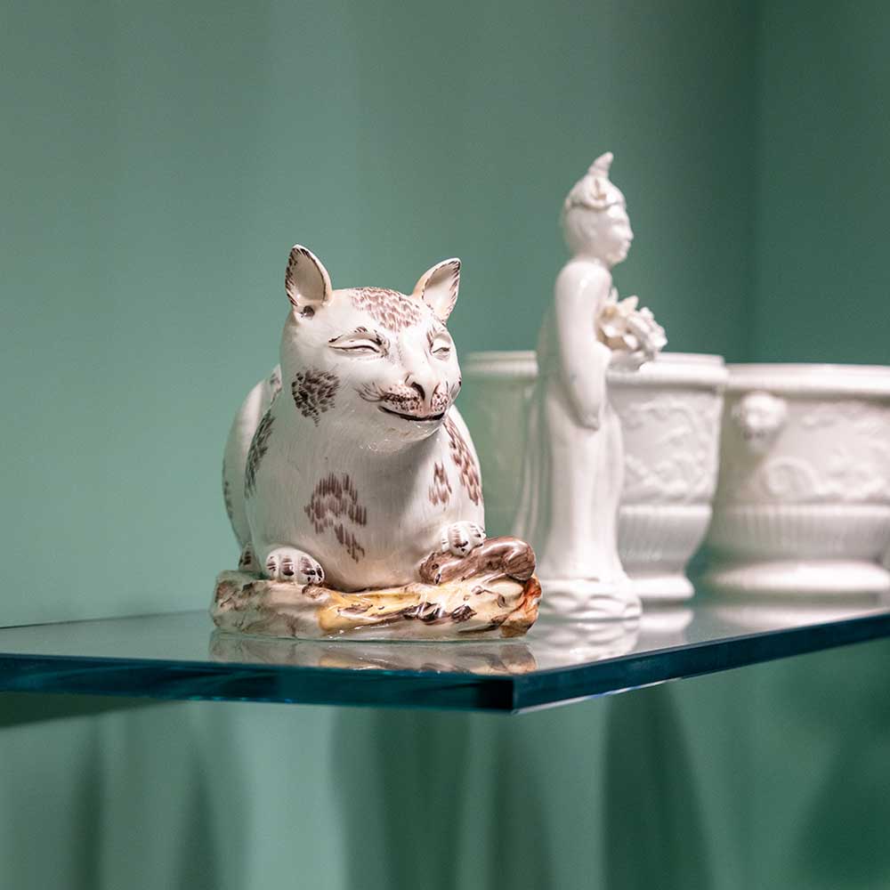 A ceramic cat and a white ceramic figure on a glass shelf with a green background