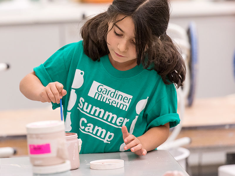 A child with pigtails and wearing a Gardiner Museum Summer Camp t-shit glazing ceramics