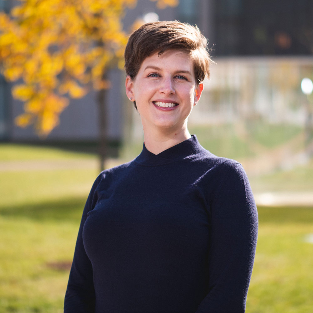 Dr. Kristina Kleutghen wearing a black shirt standing in front of grass and a tree with yellow leaves